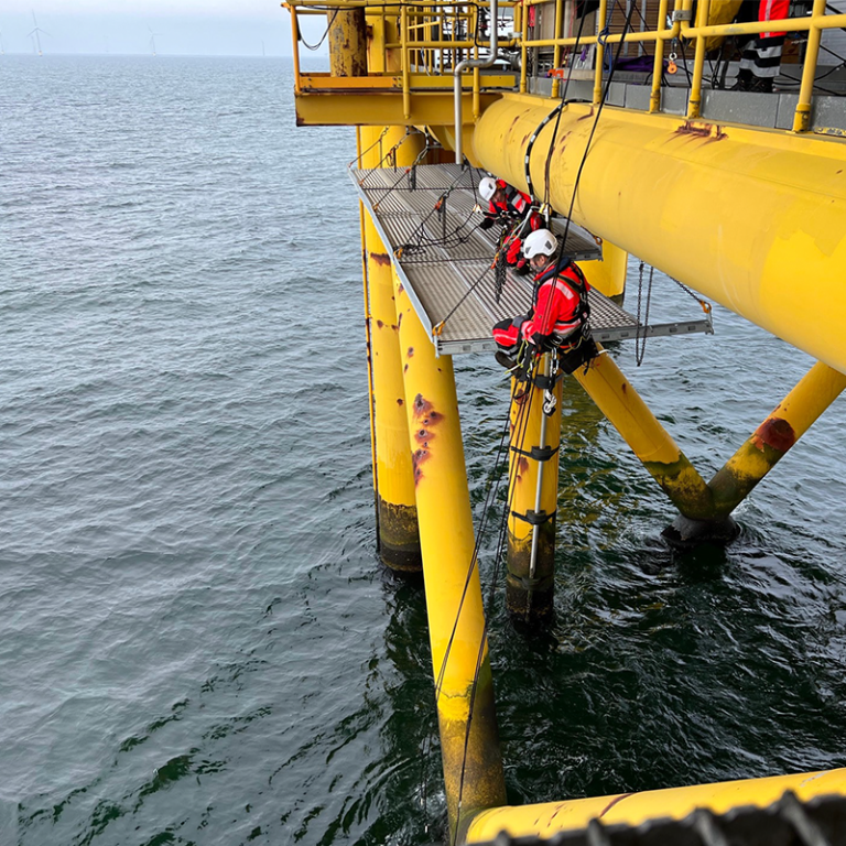 To rope access teknikere er i gang med at opsætte systemhængestilladser på jacket platform, for de kan begynde af sandblæse og male riggen.